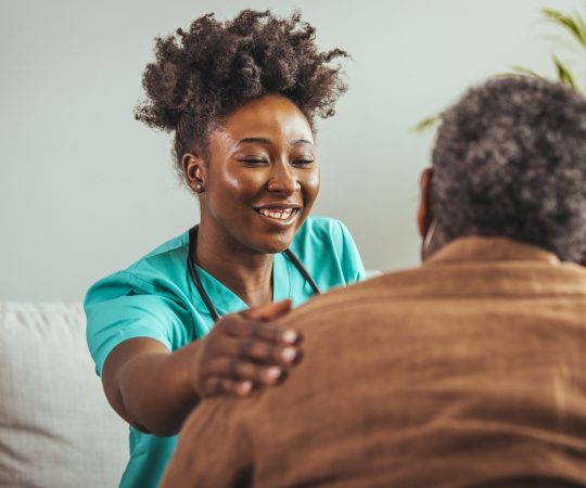 A cheerful black mid-adult female nurse attends to an elderly black male patient in a home setting, displaying professional compassion and personalized patient care.
