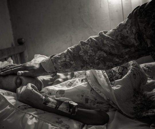 Nakiwala Halima, 31 anos, solteira, 1 filho, paciente HIV/câncer do HAU
Com Josephine Nabitaka e Ekyampaire Doreck, vamos visitar Nakiwala Halima, 31 anos, que está em cuidados paliativos. A mãe, Kivumbi Amina, 55 anos, é sua cuidadora e nos recebe muito bem. Ela agradece nossa visita e elogia o trabalho do hospice, que além do suporte emocional, fornece os medicamentos para que ela não tenha dor. Podemos ver a morfina líquida na cômoda, ao lado da cama. Ao entrarmos no quarto, André sabe que teremos pouco tempo e começa a fotografar, apesar da situação ser difícil. Halima está magra e muito mal; ela tem HIV e desenvolveu um câncer na coluna, que dificulta sua locomoção e a deixa com muita dor. Halima tem um filho de 7 anos e não é casada. Sua mãe teve 8 filhos e perdeu 3 deles. Hoje, se dedica aos cuidados de Halima e da neta, que estava na escola no momento da visita.
Quando chegamos, a mãe estava alimentando a filha. Ela está feliz em ver melhoras da filha, que agora está conseguindo se alimentar melhor e andar um pouco até o banheiro. O cuidado dessa mãe e o carinho com que cuida de Halima nos comove. Josephine e Doreck conversam com a mãe, que conta como está a filha nos últimos dias. Halima não consegue falar conosco. Ela entende nossa visita, mas está muito fraca. Seu olhar está distante. É como se estivesse longe de onde está seu corpo… Pensamos que talvez esteja mesmo perto da sua hora de partir e nos comovemos. Logo Halima se cansa e está na hora de partirmos. Na sala de visitas, fotos dos filhos de Amina. Dois deles, nos retratos, já se foram. Antes de partir, André tenta fazer uma foto da mãe e da filha (que é uma das melhores fotos do dia). Ele pensa em quantas fotos assim já fez para tantas pessoas que sabia, iam partir.
Saimos de lá um pouco tristes, mas tocados com o carinho dessa mãe. Como disse o André para ela, queríamos ter uma mãe como Amina.
 Hospice Africa Uganda, criado em 1993, para levar cuidado
